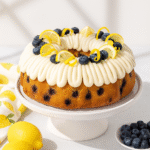 Slice of lemon blueberry Bundt cake with cream cheese frosting on a white plate, with the rest of the cake and fresh lemons and blueberries in the background.