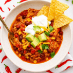 Bowl of chicken chili topped with avocado, sour cream, cheese, and cilantro, served with tortilla chips on a white counter