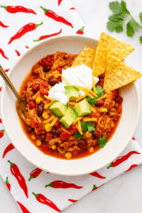 Bowl of chicken chili topped with avocado, sour cream, cheese, and cilantro, served with tortilla chips on a white counter