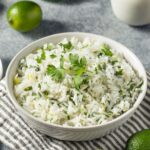 Bowl of cilantro lime rice with lime wedges and cilantro sprigs on a white surface
